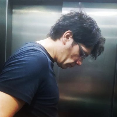 x0y0z0 Profile of a man in an elevator. He wears a blue tee-shirt, has dark hair and glasses and looks down.