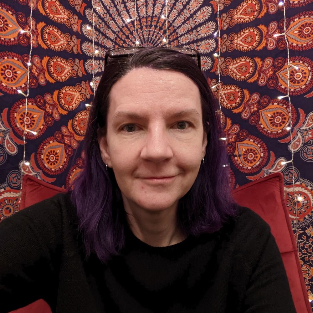 Jess Hewitt A person with purple hair wearing a black sweater sits against a vibrant, patterned backdrop, featuring lights and a cozy red chair.