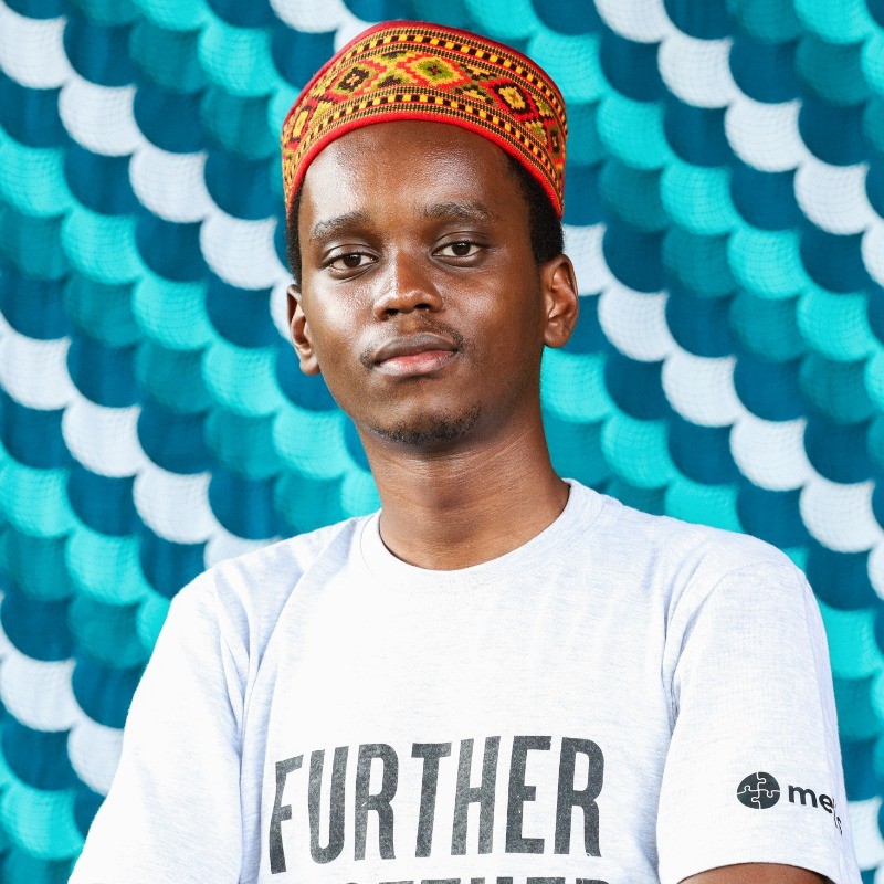 A person stands confidently in front of a vibrant, textured blue backdrop, wearing a colorful traditional hat and a white shirt with "FURTHER" printed on it.