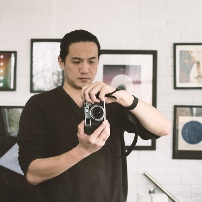Wen Dark haired man in a brown shirt holding a camera sideways. Pictures on a wall behind him.